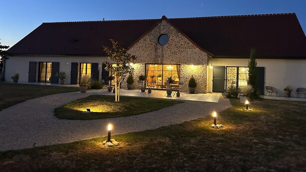 A stone building with a red roof, large windows, and a well-lit outdoor seating area. - Aux 3 Forets (Bizeneuille)