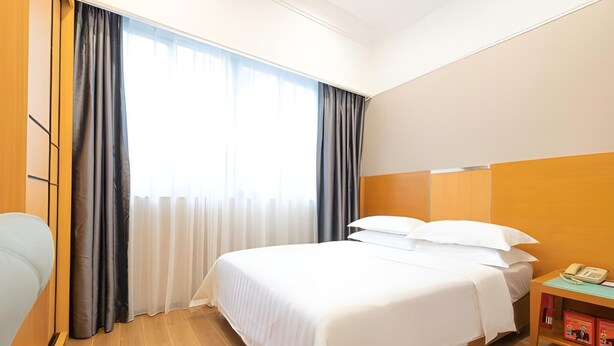 A hotel room with a large bed, a wooden headboard, a bedside table with a phone, and a window with curtains. - Carrianna (Foshan)