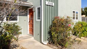 Un edificio residencial con fachada verde, un cartel con el número 2048, una puerta de madera y un pequeño jardín con diversas plantas y flores.