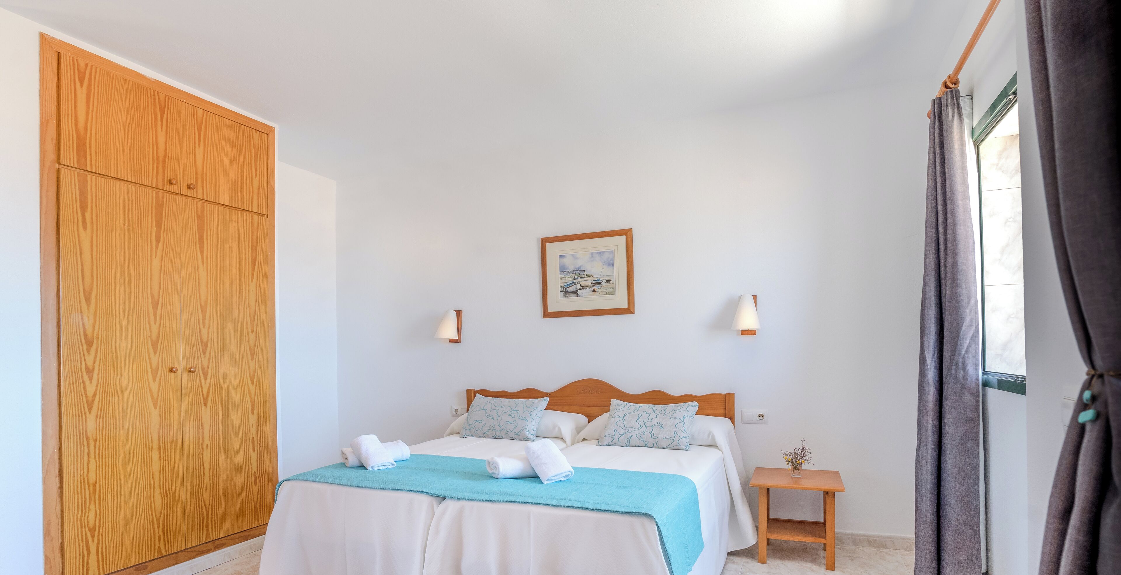 Ein Schlafzimmer mit Holzschrank, einem Bett mit weißer Bettwäsche und einer blauen Decke, einem kleinen Nachttisch mit Lampe und einem Fenster mit Vorhängen.