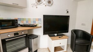 Eine kompakte Küche mit Mikrowelle, Herd und Toaster. Ein Flachbildfernseher ist an der Wand befestigt, und ein schwarzer Stuhl steht daneben.