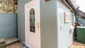 Ein kleines, eingeschossiges Gebäude mit einer weißen Tür und einem Fenster mit dekorativer Glasverkleidung. Links ist ein Metallgitter und rechts eine Satellitenschüssel zu sehen.