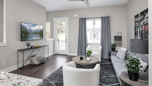 A modern living room with a flat-screen TV, a ceiling fan, a sofa, a coffee table, and a large window with curtains. - Nashville Luxe Retreat • Rooftop & Hot Tub (Nashville)