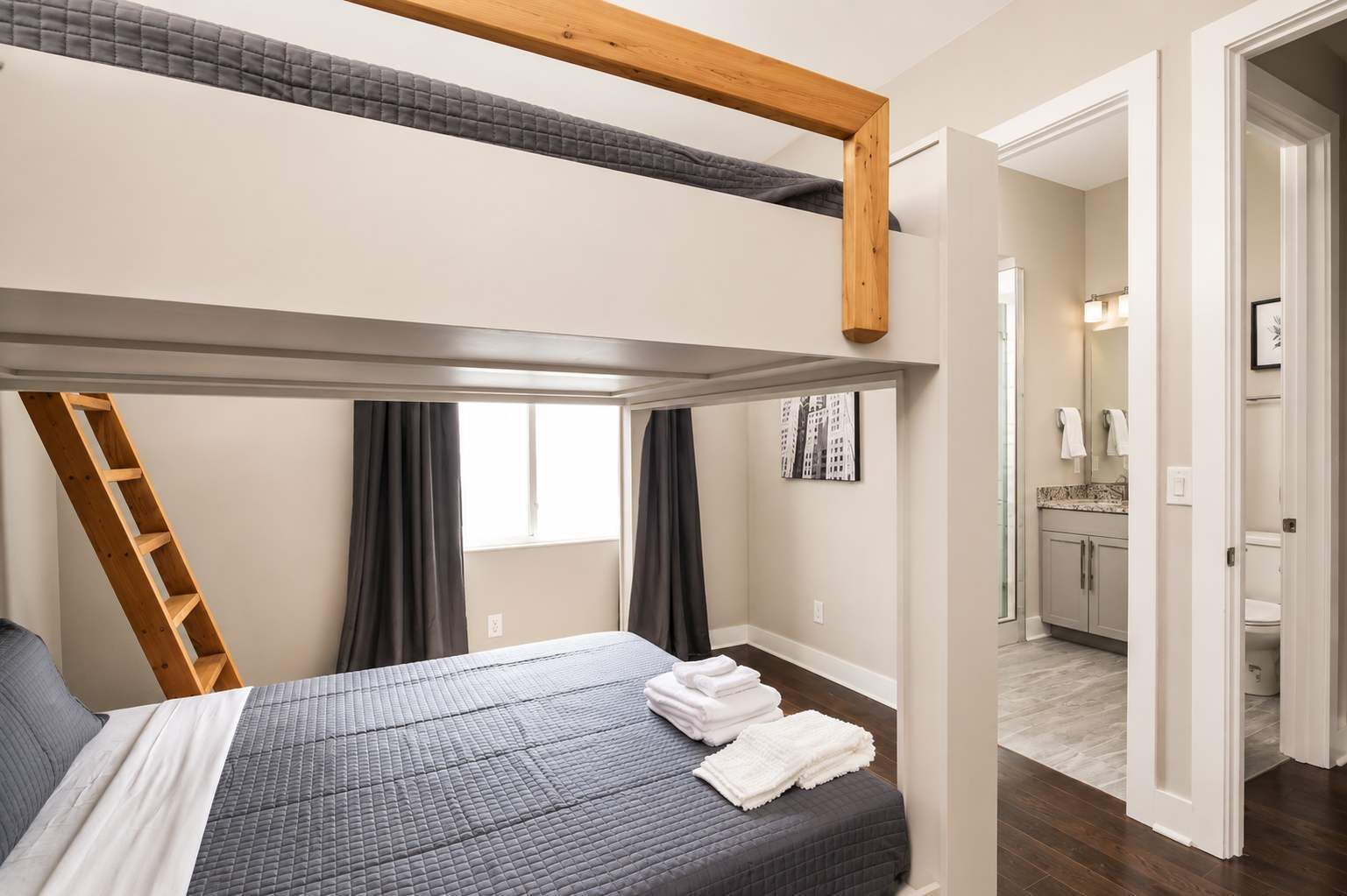 Una camera da letto con un letto a castello, una scala in legno, una finestra con tende e un bagno parzialmente visibile attraverso una porta aperta.