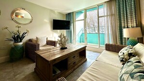 Un salon avec une table basse en bois, un canapé, un téléviseur fixé au mur et une grande fenêtre avec des rideaux.