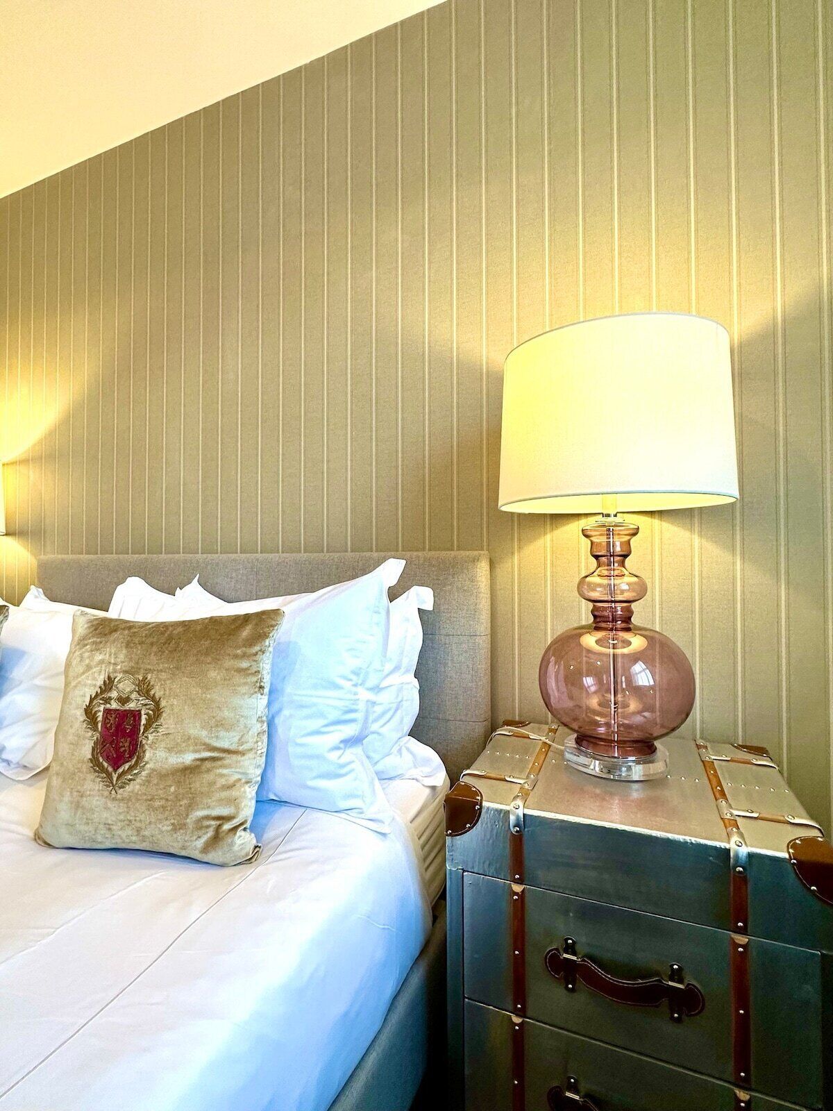 Un lit bien fait, avec des oreillers blancs et bleu clair, un coussin décoratif orné d’un blason, et une lampe de style vintage posée sur une table de chevet en métal.