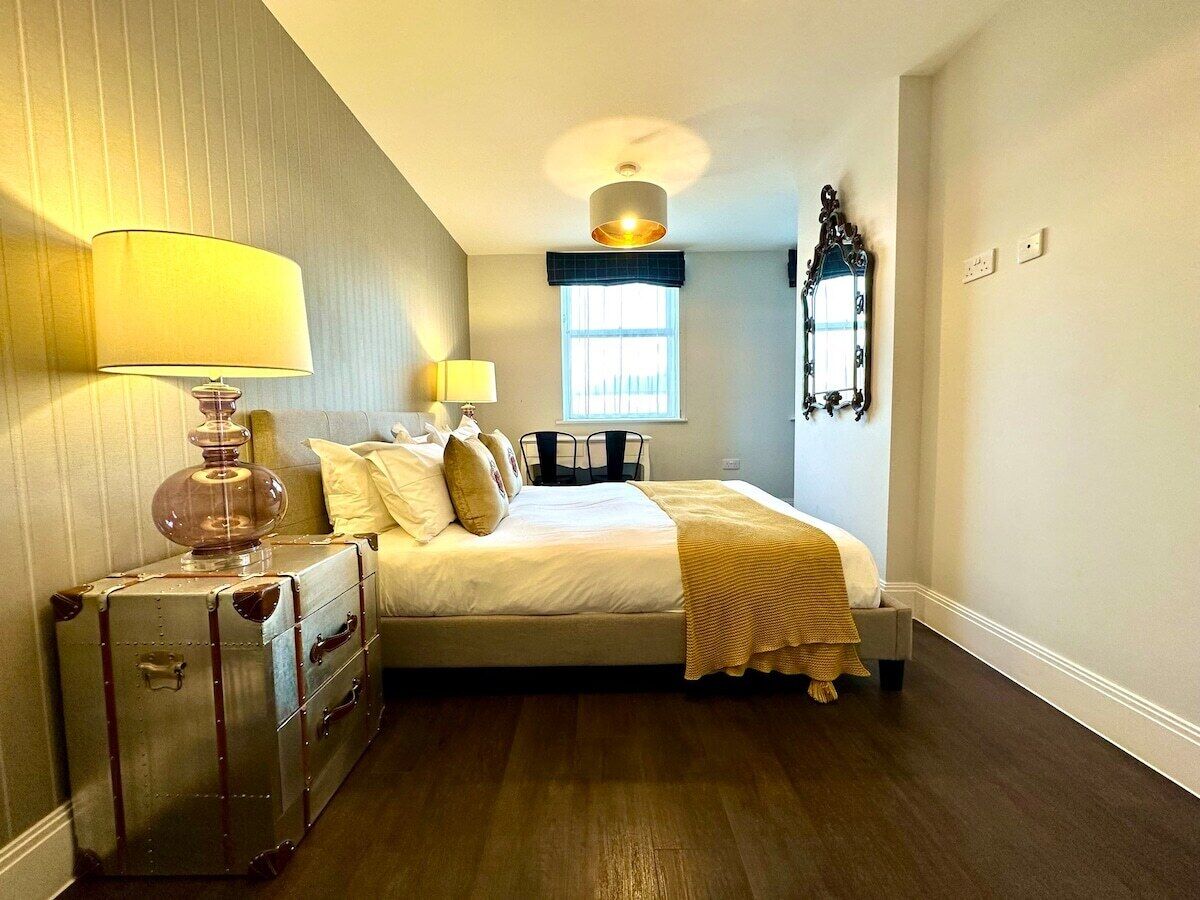 A bedroom with a bed, two bedside lamps, a window, a mirror, and a vintage suitcase.