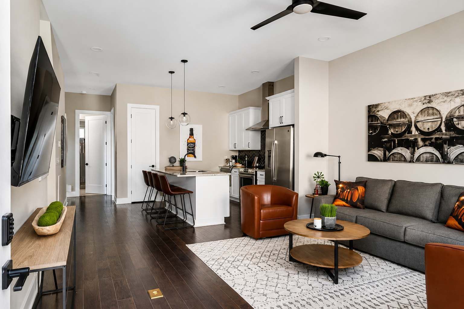 A modern kitchen with a central island, a dining area with bar stools, a living room with a sofa and coffee table, and a ceiling fan.