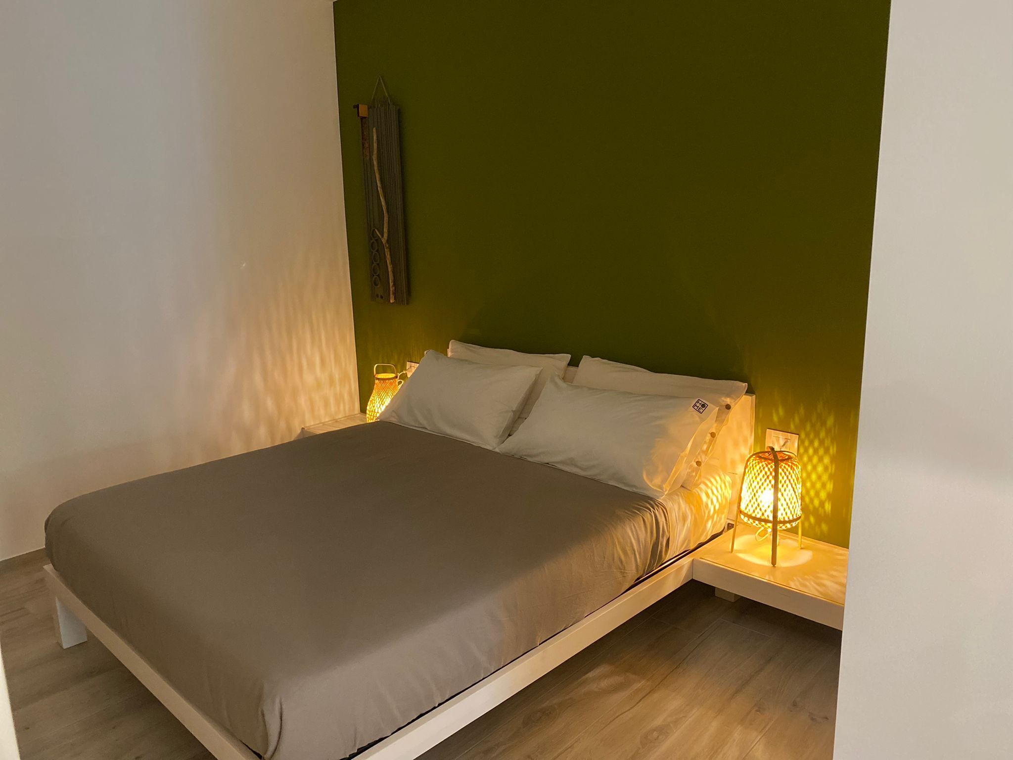 Una cama individual con colchón gris y almohadas blancas, una mesita de noche con lámpara, y una pared verde.