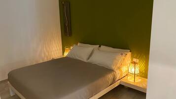 Una cama individual con colchón gris y almohadas blancas, una mesita de noche con lámpara, y una pared verde.