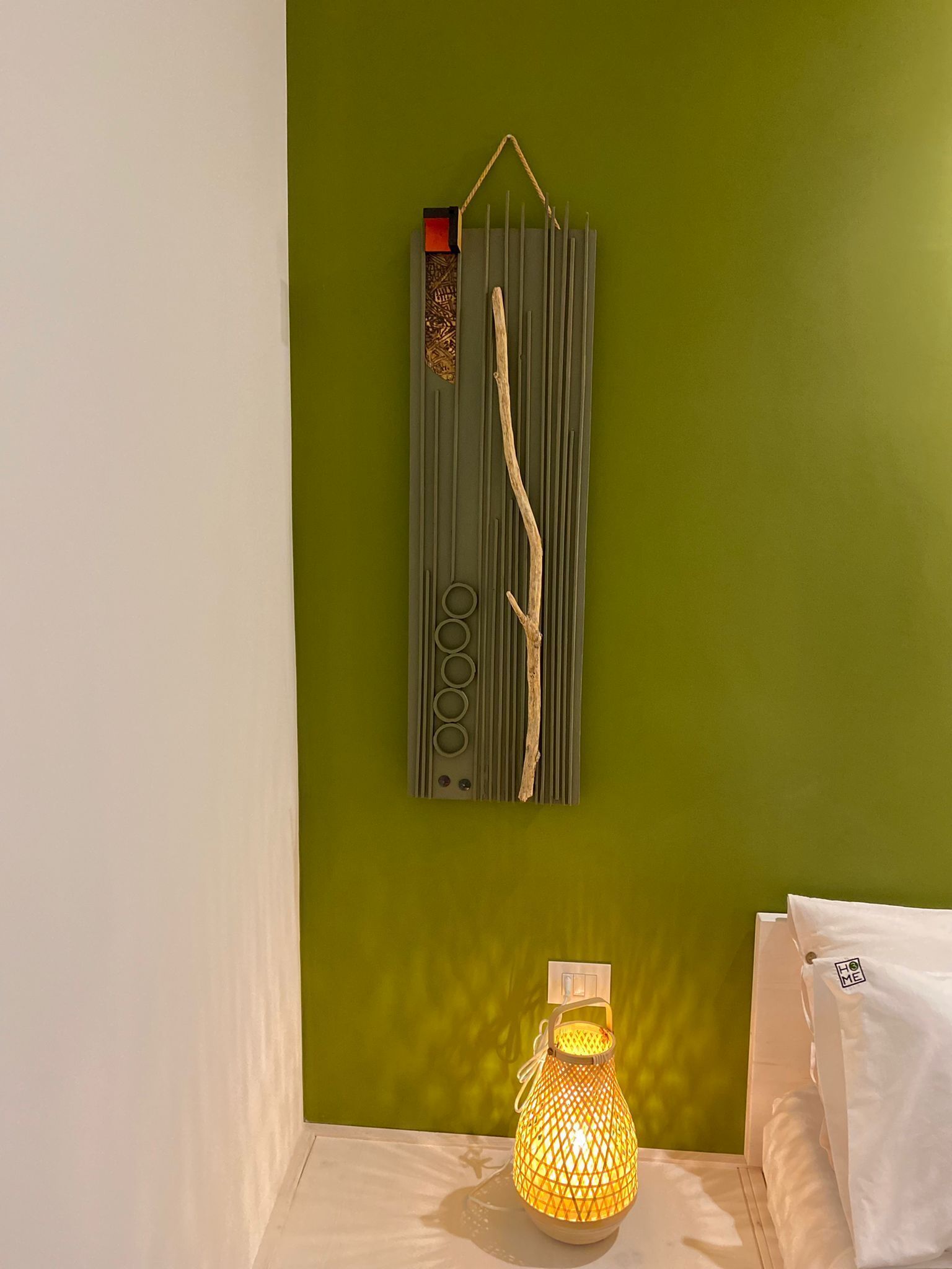 Ein Zimmer mit grüner Wand, einem an der Wand befestigten dekorativen Element mit vertikalen Linien und einem Zweig, sowie einem Tisch mit einer beleuchteten Laterne.