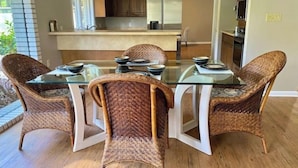 A dining area with a glass table surrounded by wicker chairs, a kitchen in the background, and a wooden floor. - Oak Rock Ranch - Riverfront - Scenic Old Florida (Inglis)