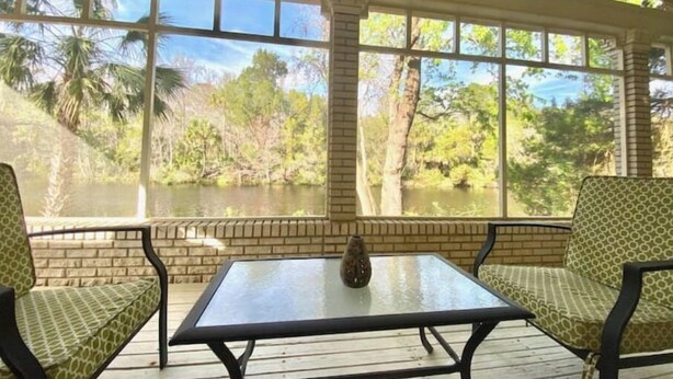 A covered patio with a glass table and two patterned chairs, overlooking a natural landscape with trees and a body of water. - Oak Rock Ranch - Riverfront - Scenic Old Florida (Inglis)