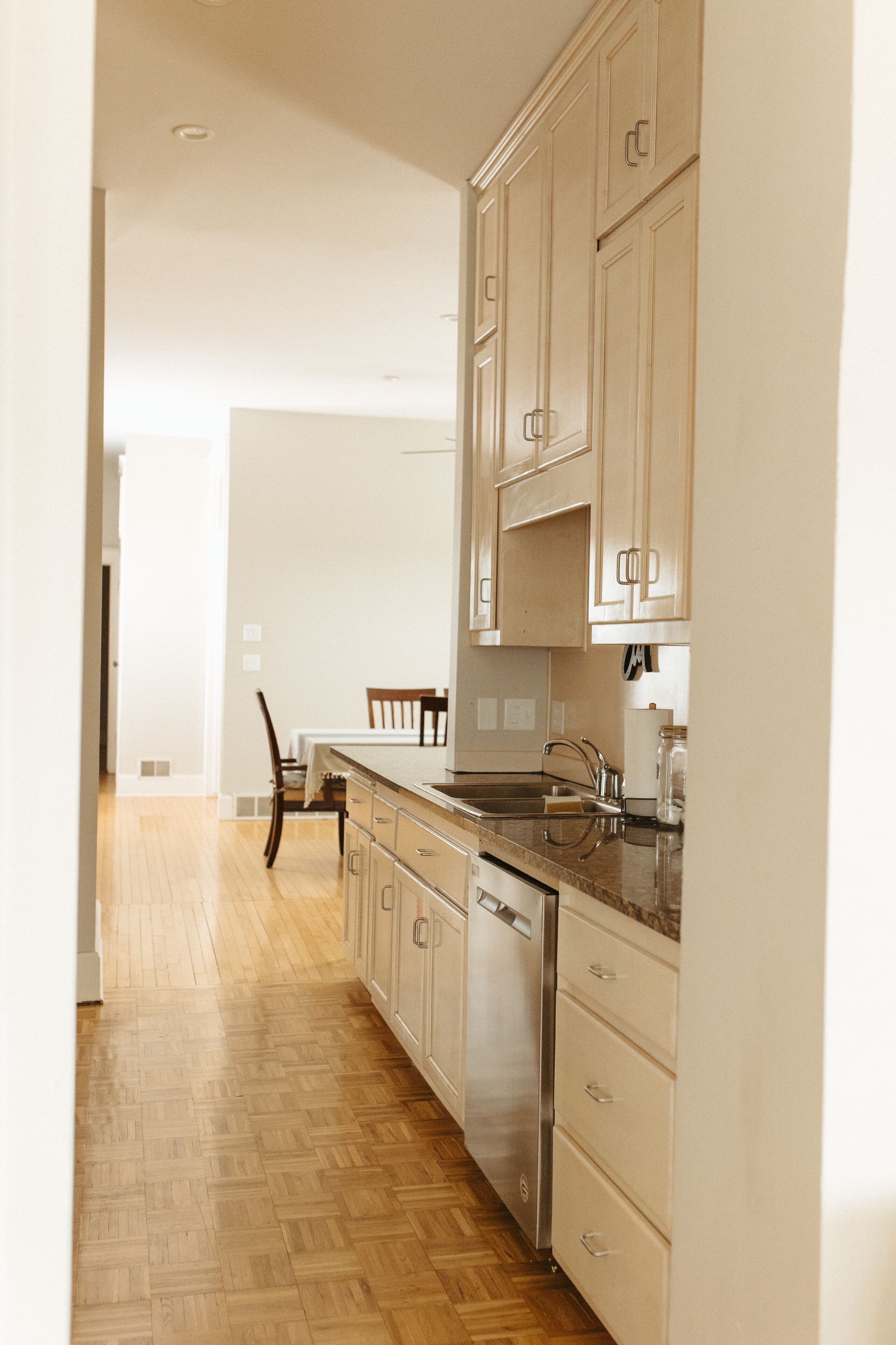 Une cuisine avec un plancher en bois, des armoires de couleur beige, un lave-vaisselle et un coin repas comprenant une chaise et une table.