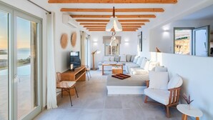 A modern living room with a large sofa set, a coffee table, and a TV. There is a wooden ceiling with exposed beams, and a large window offering a view of the sea. - Villa Flo by Anamnesia Collection (Naxos)