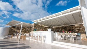 An outdoor patio with a stone wall backdrop, white furniture, and a pergola structure. - Villa Flo by Anamnesia Collection (Naxos)