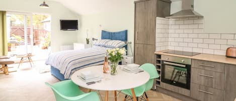 Quarto moderno com uma cama grande, uma pequena mesa de jantar com cadeiras, área de cozinha com fogão e armários, e uma TV fixada na parede.