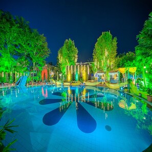 A swimming pool with a slide and a small waterfall feature, surrounded by trees and illuminated by colorful lights. - Renest Shraddha Inn Shirdi (Rahata)
