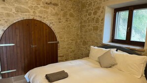 Ein Schlafzimmer mit Stehwand, Holztür, einem Bett mit weißer Bettwäsche und einem Fenster mit Blick auf Bäume.