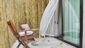 A wooden chair with cushions, a small table, and a white curtain in a room with a bamboo wall and a glass door.