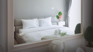 A modern hotel room with a large bed, a bedside table with a lamp, a small potted plant, and a view of the outside through a window with curtains.