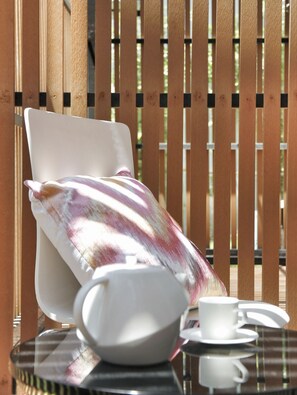 A sunlit outdoor seating area with a cushioned chair, a table with a teapot, cups, and saucers.