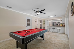 A pool table in a room with a ceiling fan, a kitchen area with a refrigerator and microwave, and a door leading to another room. - Spacious & Amazing Home w/ Big Pool and Patio (Brandon)