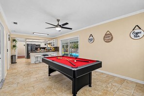 A pool table in a room with a ceiling fan, a kitchen area, and a pool visible through a glass door. - Spacious & Amazing Home w/ Big Pool and Patio (Brandon)