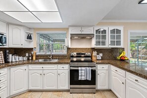 A kitchen with white cabinetry, a stainless steel oven, a granite countertop, and a window with a view of greenery. - Spacious & Amazing Home w/ Big Pool and Patio (Brandon)