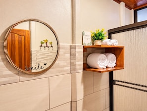 A bathroom with a round mirror, a wooden shelf with towels, and a decorative sign. - Arenal Dream Cottage (La Fortuna)