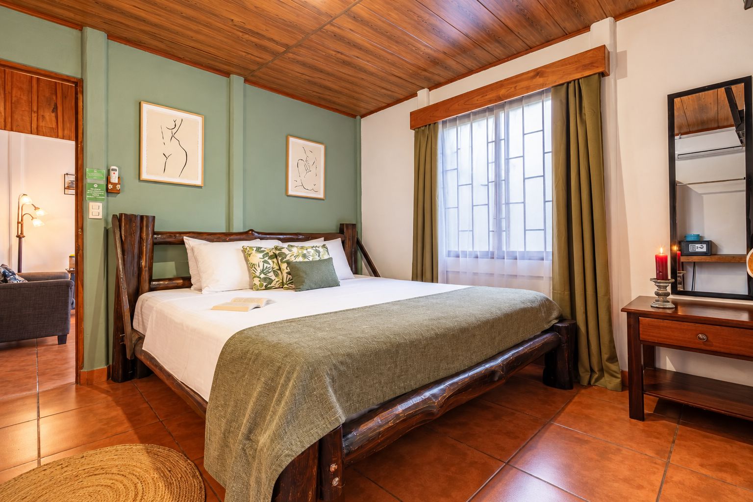 A hotel room with a wooden bed, a bedside table, a mirror, and a window with curtains.