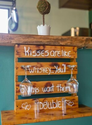 Ein hölzernes Schild mit einer humorvollen Aufschrift über Küsse und Whiskey, dazu zwei leere Gläser.