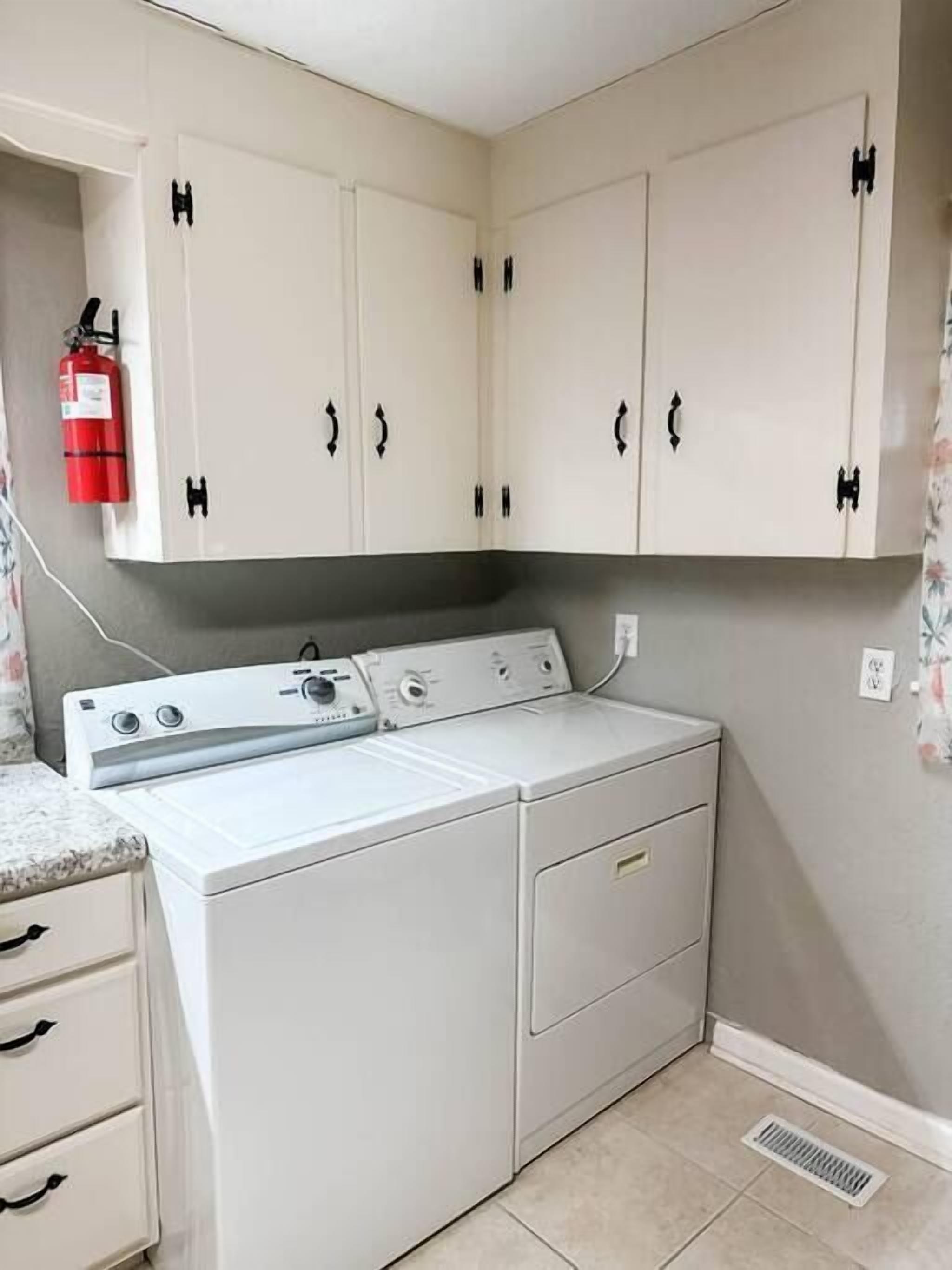 A laundry room with a front-loading washer and dryer, beige cabinets with black handles, a fire extinguisher on the wall, and a power outlet.
