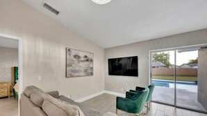 A living room with a beige sofa, a TV mounted on the wall, and a sliding glass door leading to a backyard. - Casa De Paradiso - Modern yet comfy home at the center of it all! (Kissimmee)