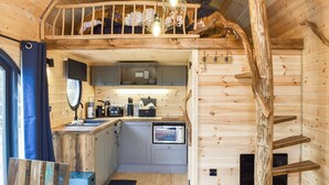 Interior d'una cabana de fusta amb cuina, zona de menjador i un dormitori en alt amb un llit i una làmpada penjant.