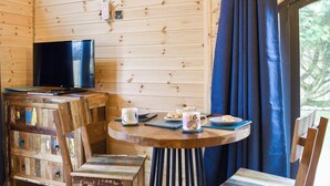 Interior acollidor d'una cabana amb elements de fusta, una televisió sobre un moble de fusta, una taula rodona amb una tassa i galetes, i una vista per una finestra amb cortines blaves.