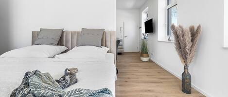 Ein modernes Schlafzimmer mit Bett, Kissen und Decke. Ein Flur führt zu einem Badezimmer mit Dusche und Fenster.