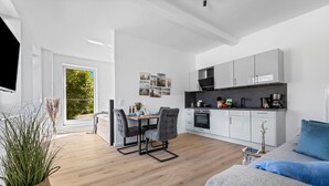 Een moderne keuken met witte kasten, een zwart aanrecht en een koffieapparaat. Er is een eethoek met een tafel en stoelen, en een groot raam met uitzicht op groen buiten.