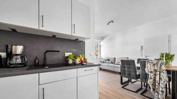 A modern kitchen with white cabinets, a black countertop, and a coffee machine. There is a dining area with a gray sofa and a table with chairs.