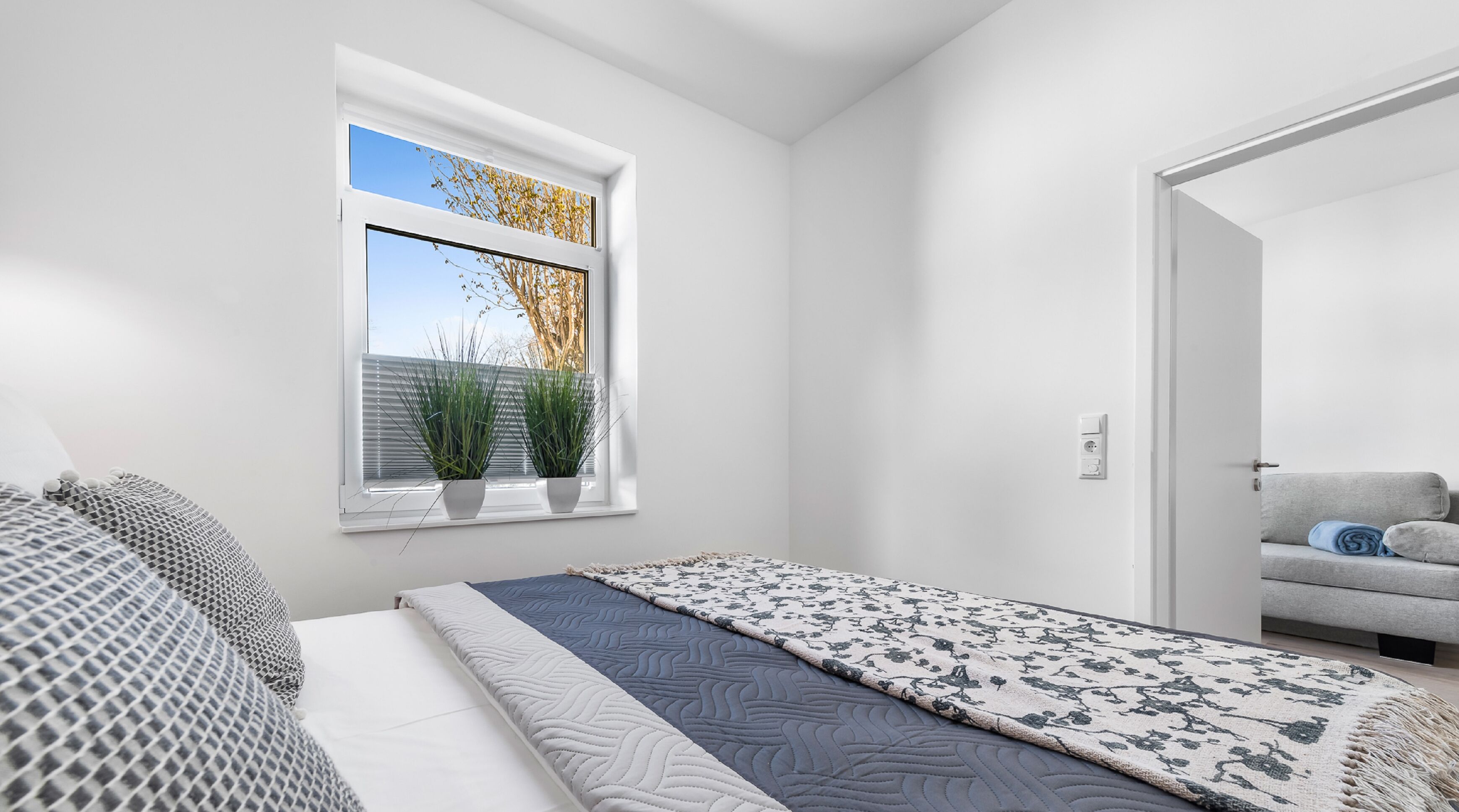 Un dormitorio moderno con una cama grande, una ventana con persianas, dos plantas en macetas y un sofá gris en la habitación contigua.