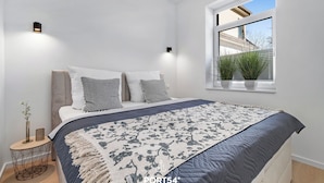 A modern bedroom with a large bed, a window with plants, and a geometric side table. - Strandläufer - Shower, TV, Terrace, Garden Furniture, Grill, Parking Space (Gelting)