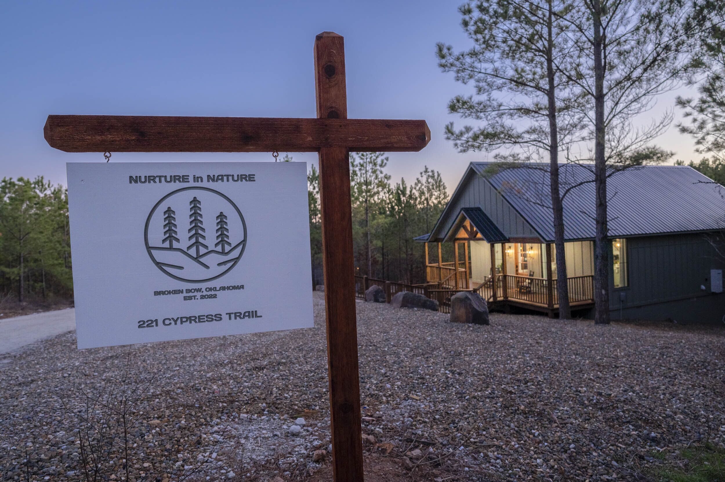 A wooden signpost with a white sign for 'Nurture in Nature' at 221 Cypress Trail, a rustic cabin with a porch, and a gravel path.