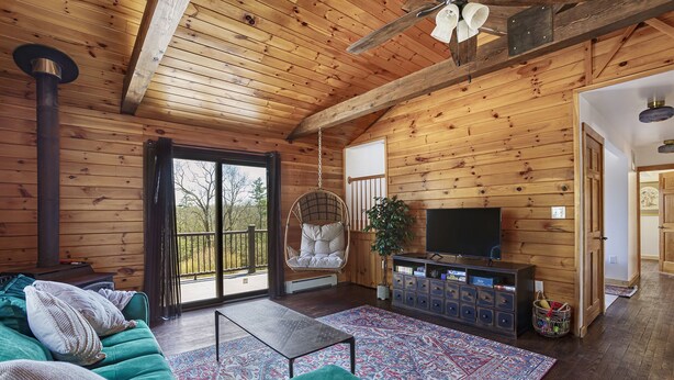 Interior acogedor de una cabaña con techo de madera, un ventanal grande, un sofá verde, un televisor y una silla colgante.