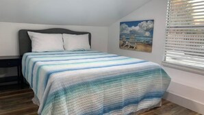 Ein Schlafzimmer mit Bett, Nachttisch, einem Bild von einer Strandlandschaft und einem Fenster mit Jalousien.