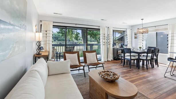 Una sala de estar moderna con sofá, mesa de comedor y sillas, ventanas amplias con cortinas y piso de madera.