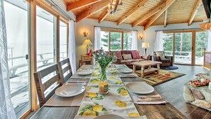 Un comedor con una mesa larga dispuesta para cuatro personas, vigas de madera en el techo y grandes ventanales que ofrecen vistas al exterior.