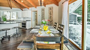 Un comedor con una mesa de madera dispuesta para cuatro, una cocina al fondo y un gran ventanal con vistas a un paisaje nevado.