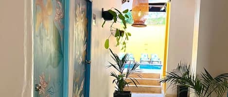 Un pasillo con dos puertas, cada una decorada con motivos florales, que conducen a una habitación. Hay plantas en macetas a lo largo del pasillo y una lámpara colgante arriba.