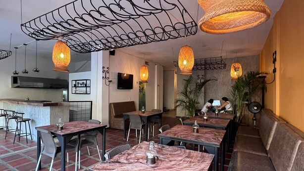 Un restaurante con mesas y bancos de mármol, lámparas de mimbre colgantes y una zona de cocina al fondo.