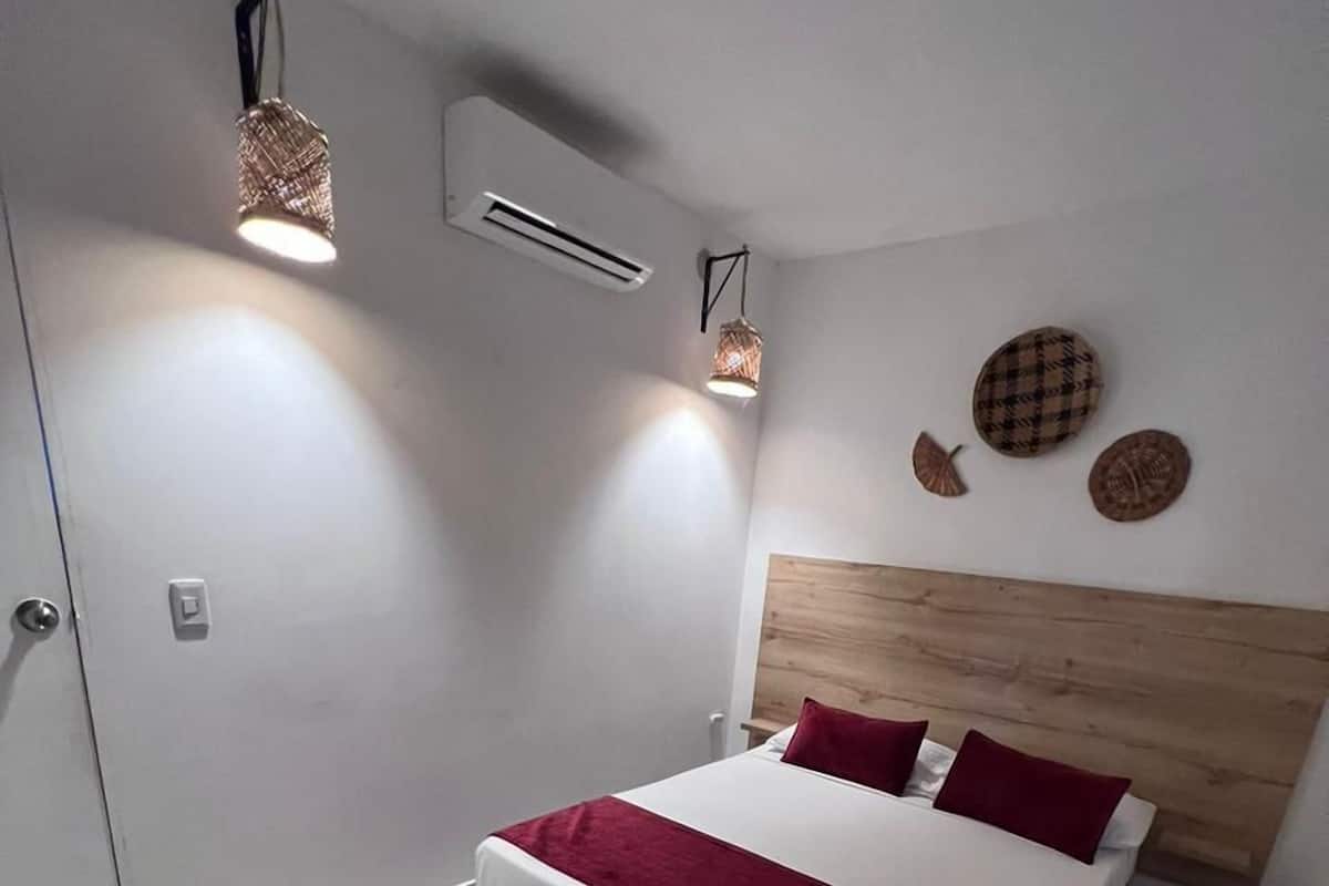 Un dormitorio moderno con cabecera de madera, una cama grande con ropa de cama roja y blanca, dos lámparas colgantes, un equipo de aire acondicionado y una pequeña mesa con una silla.